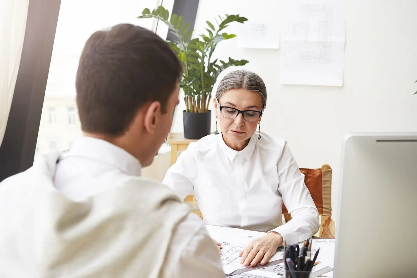 Изображение стильной 50‑летней женщины — главного архитектора в очках, проверяющей чертежи неузнаваемого сотрудника-брюнета, изучающей документацию на столе, с серьезным сосредоточенным выражением лица.