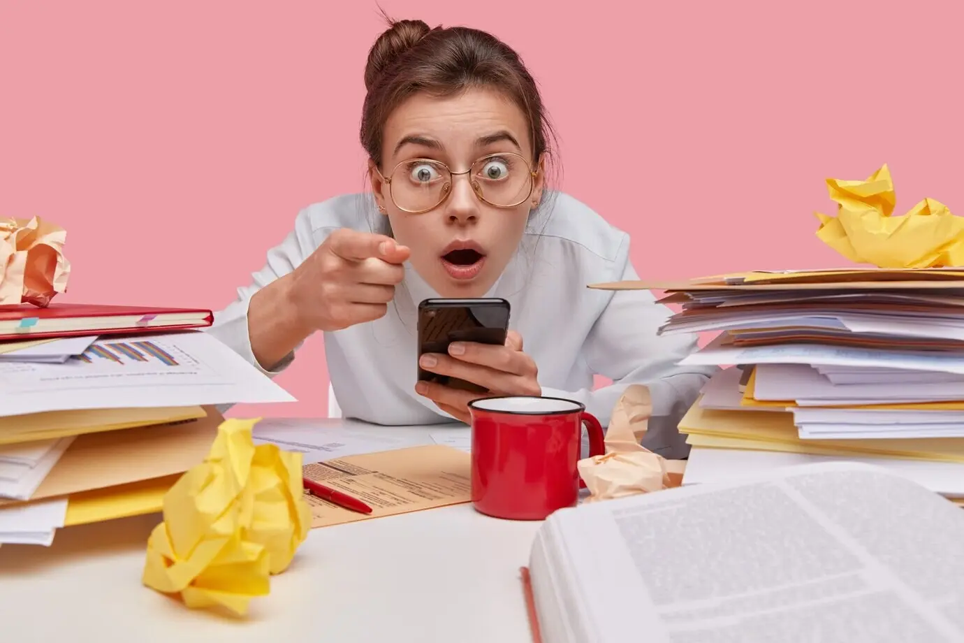 Снимок в помещении: шокированная молодая европейская женщина держит современный мобильный телефон, в ступоре указывает на камеру, в больших очках, с открытым ртом.